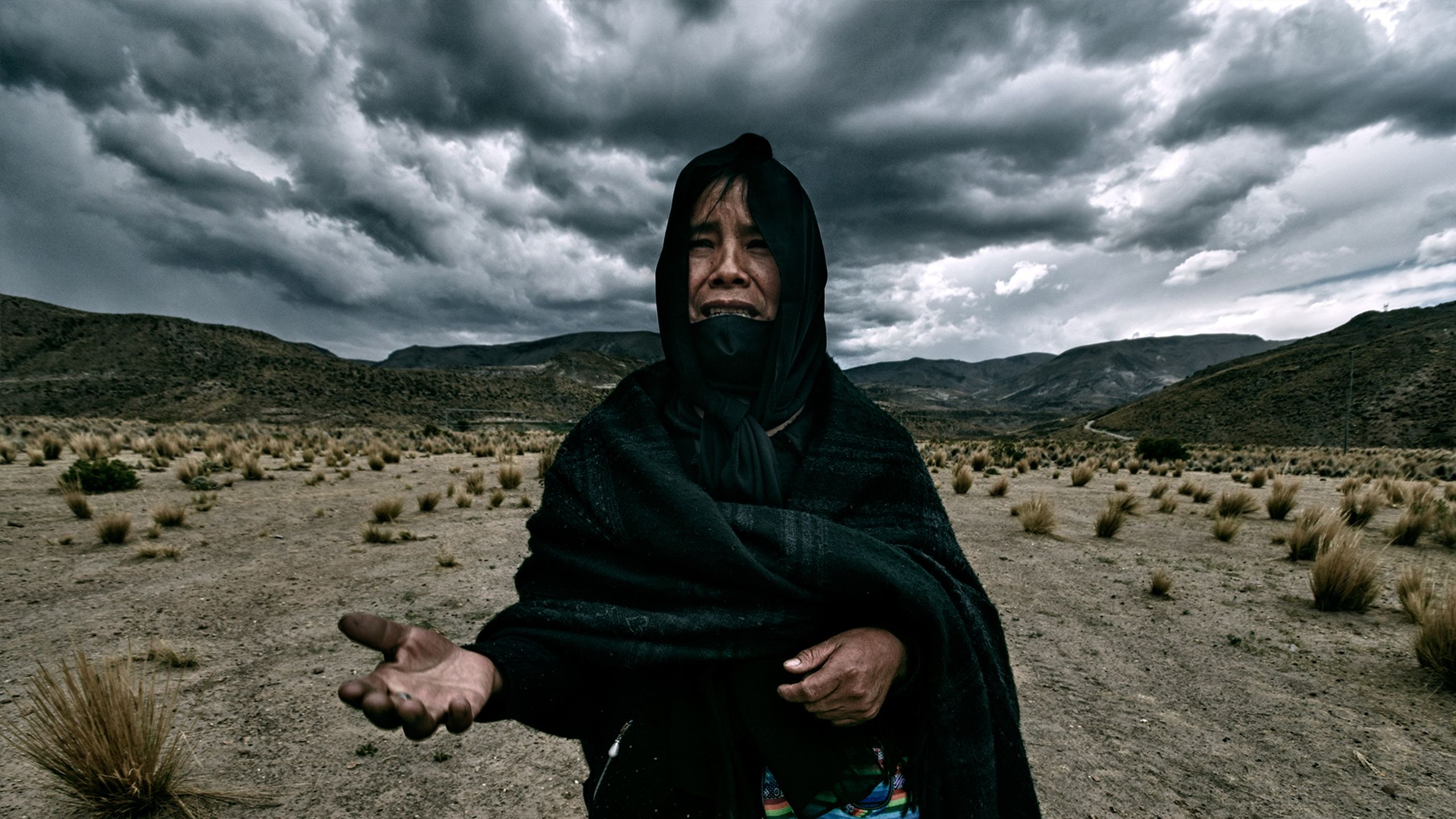 A Bolivian woman in traditional mourning dress stretches out her hands accusingly before a cloudy sky and barren landscape.