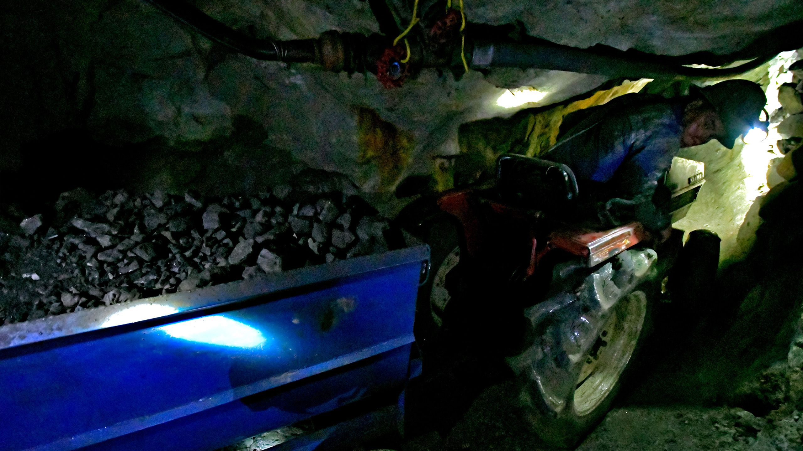 Un ouvrier conduit un petit tracteur et une remorque pleine de minerais dans une galerie étroite. Le tunnel est si bas que le conducteur doit se baisser pour passer.